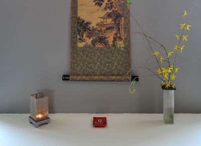 Counter with candle, incense, and forsythia floral arrangement. Part of a scroll visible on wall.